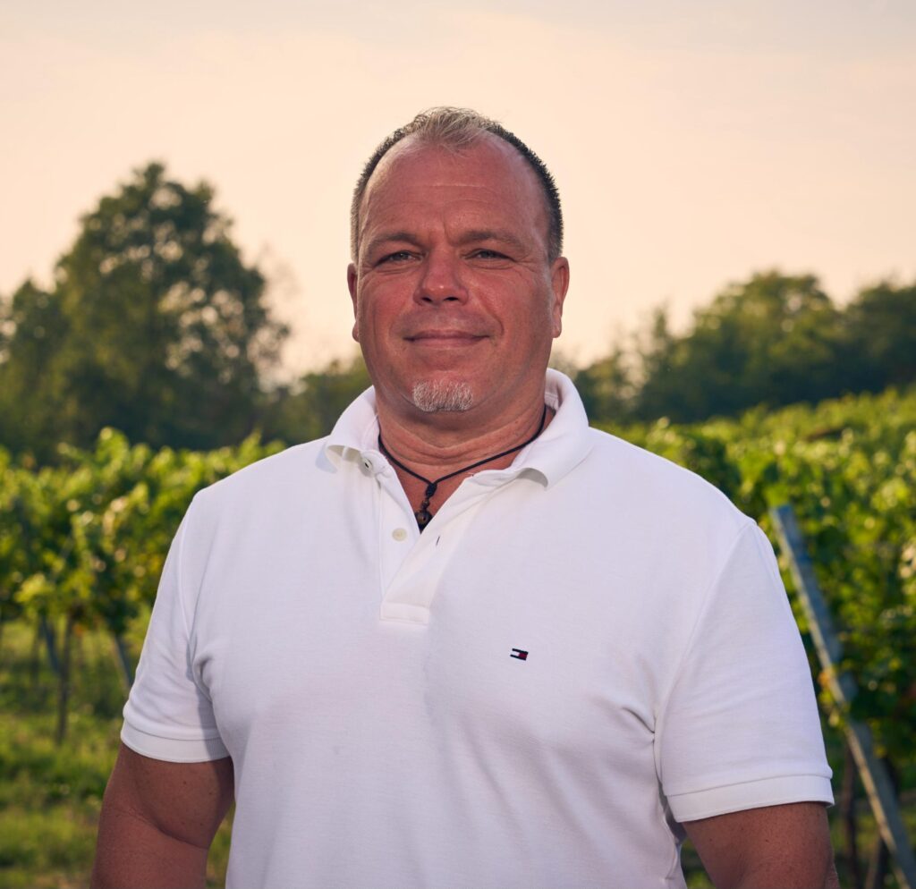 Man in a white polo shirt outdoors with trees and greenery in the background.