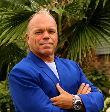 Portrait of a man in a blue blazer with arms crossed, standing outdoors among palm leaves.